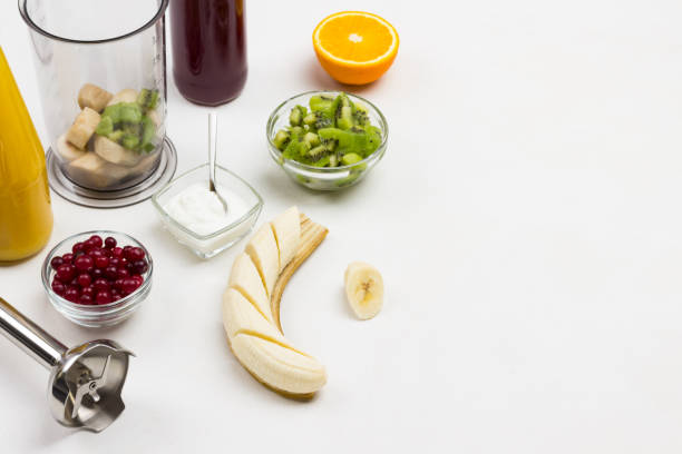 Sliced banana and peel. Half orange. Cranberries and kiwi in bowls. Blender and banana blender jar. Ingredients for making fruit smoothies. White background. Top view. Copy space