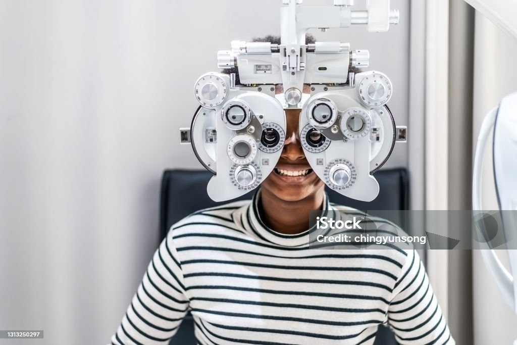 Smiling young woman african american afro hair doing Eye test on optical phoropter, checking on her eye with optometry machine Eye Care Professional Stock Photo