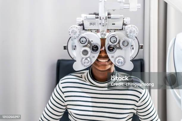 Smiling Young Woman African American Afro Hair Doing Eye Test On Optical Phoropter Checking On Her Eye With Optometry Machine Stock Photo - Download Image Now