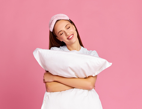 Cheerful young blond woman hugging pillow closed eyes with smile while dreaming about sleep against pink background