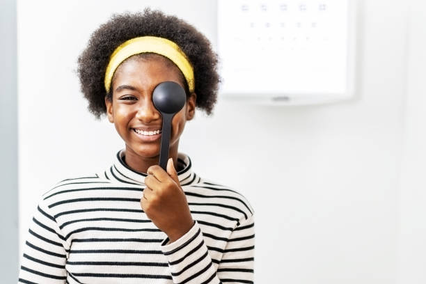 jeune femme africaine de sourire faisant l’essai d’oeil dans les cliniques ophtalmologiques, retenant l’occluder et regardant le diagramme, correction de vue, clinique d’optique - examen ophtalmologique photos et images de collection