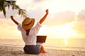 Rear view of young female freelancer in straw hat celebrating success. while working remotely on the tropical sandy beach at sunset