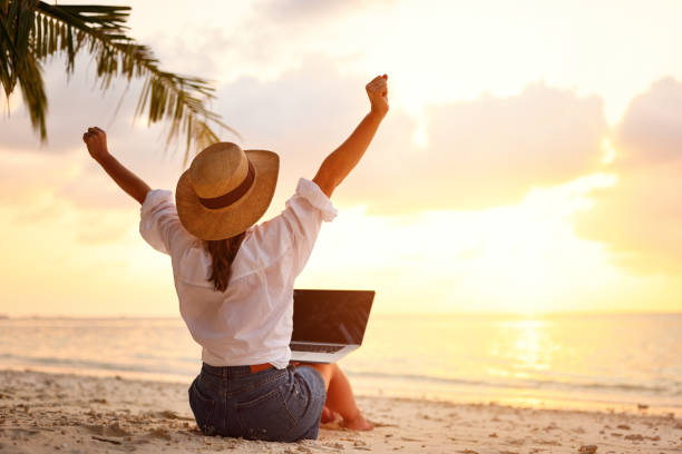 Rear view of young female freelancer in straw hat celebrating success. while working remotely on the tropical sandy beach at sunset Work from anywhere. Rear view of young woman, female freelancer in straw hat working on laptop, keeping arms raised and cellebrating success while sitting on the tropical sandy beach at sunset telecommuting stock pictures, royalty-free photos & images
