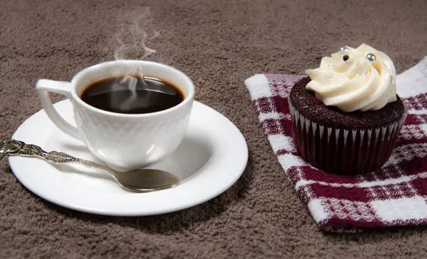 Steaming white coffee cup and homemade cupcake on brown fabric background, homemade cake with cream
