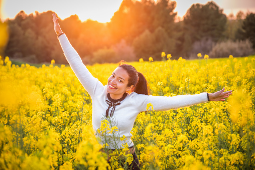 Beautiful happy caucasian brunette young woman smiling with arms outstretched in the middle of a field of yellow rapeseed flowers at sunset in spring. Soft and sensual beauty. Catalonia, spain