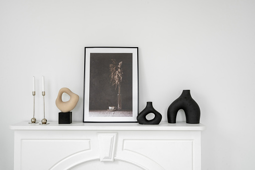 Mockup picture frame, vase and candle in candlestick standing on shelf above vintage fireplace. Fashionable living room with modern interior design and white copy space wall on background