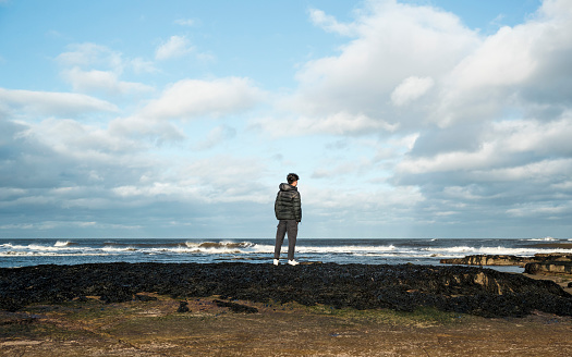 Rear view of a mixed race teenage boy standing on the beach looking out at the ocean in the North East of England. He is standing looking at the sea view in deep thought.