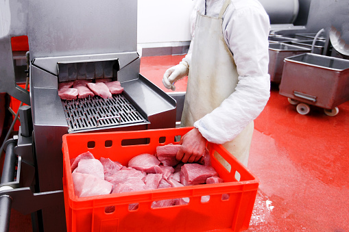 Worker standing near red plastic boxes full with raw cuts of minced meat, introduced into an introductory washing in the meat production process.