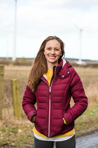 Front view portrait of a caucasian mid adult woman standing in a non urban area with wind turbines behind her looking at the camera smiling.