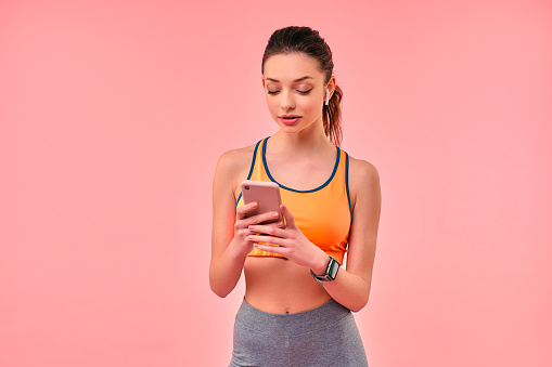 Cute sporty girl in sportswear with phone and wireless headphones isolated on pink background. Sports, yoga, active lifestyle, exercise for health.