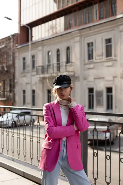 Young beautiful girl in fashionable clothes and a black hat on the street. Model in a pink jacket with rings on her fingers against the background of the city.