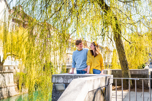 Young cheerful heterosexual couple standing under the tree by the river in a city and enjoying a view.