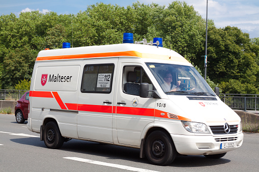 Ambulance of German Malteser healthcare is driving on Bundesstrasse in Essen Werden in summer. One man is sitting in car. Blue lights are active.