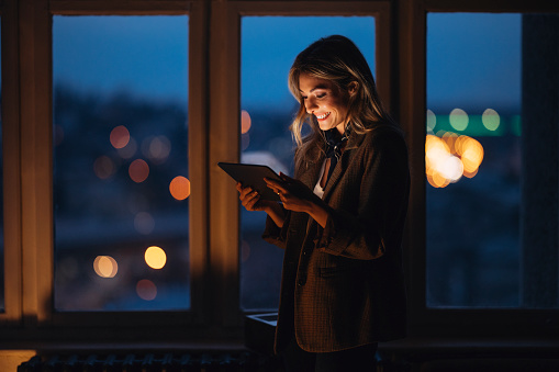 Side view of smiling female entrepreneur using digital tablet at dusk. Young businesswoman is standing by window in dark. She is in creative office.