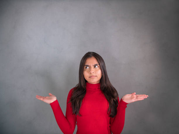 istock Young woman in red pullover, the elbows bent, arms raised and palms up. The face expression is serious, uncertain and slightly questingly. She gazes to the gray background. 1313301852