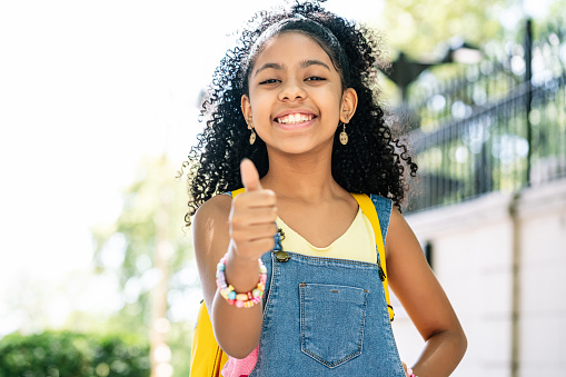 African American little girl smiling and with thumb up while wearing a backpack on the street. Education concept.