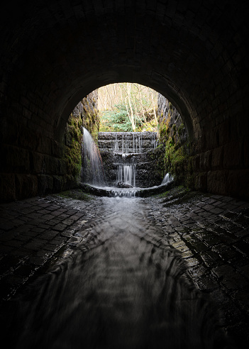 Very old under the road creepy dark eerie old sewer brick tunnel spooky shadows water river running through flowing light at the end. Waterfalls long exposure blurred flow. Atmospheric sinister culvert old underpass.