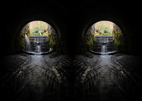 Shaded creepy dark eerie old double brick tunnels with spooky shadows water river running through flowing light at the end. Waterfalls long exposure blurred flow. Atmospheric sinister culverts old underpass.