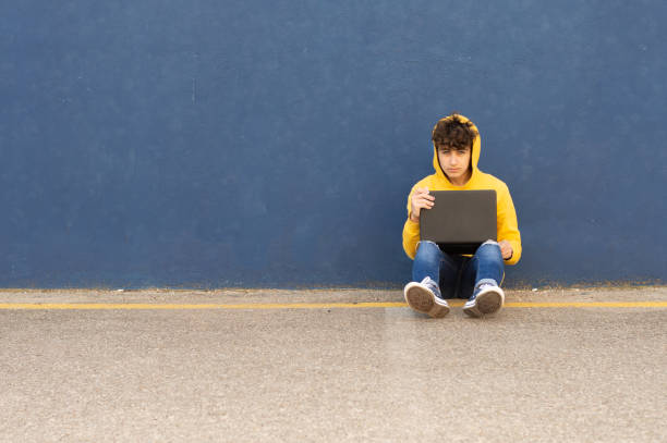Young man in yellow sweatshirt leaning against blue wall while studying with laptop. Looking at the camera. Space to copy