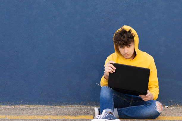 istock Young man in yellow sweatshirt leaning against blue wall while studying with laptop. Looking at the camera. Space to copy 1313311246