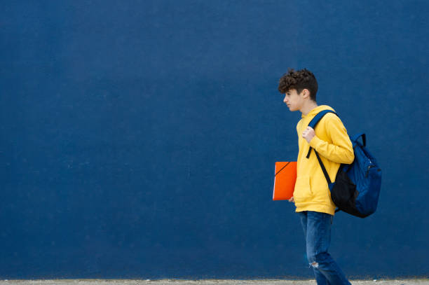 young adult student walking in yellow sweatshirt, carrying a blue backpack and orange folder. Blue wall behind. Space to copy.
