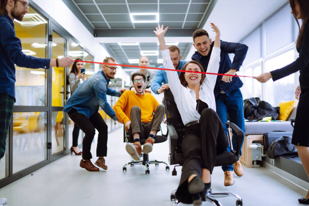 Friendly work team  ride chairs in office room cheerfully excited diverse employees laugh while enjoying fun work break activities, creative friendly workers play a game together.
