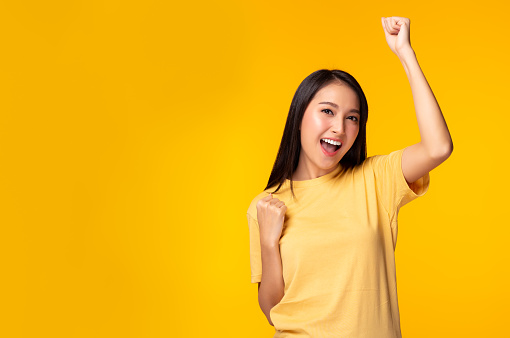 Happy excited young asian female with raised hands celebrating success Portrait cheerful asian woman with raised fists and smile face Attractive girl get happy, enjoy life yellow background copy space