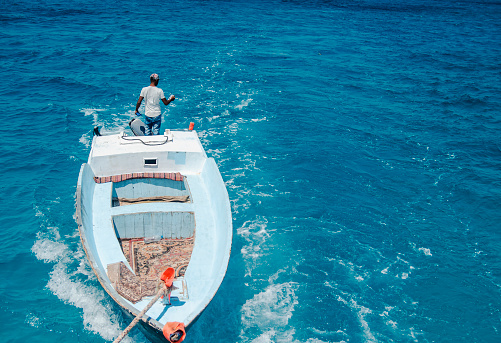 Back of Man standing in small fishing boat in the blue sea. Black Arabian fisherman sailing and looking in the water searching for the fish. Above view. Copy space