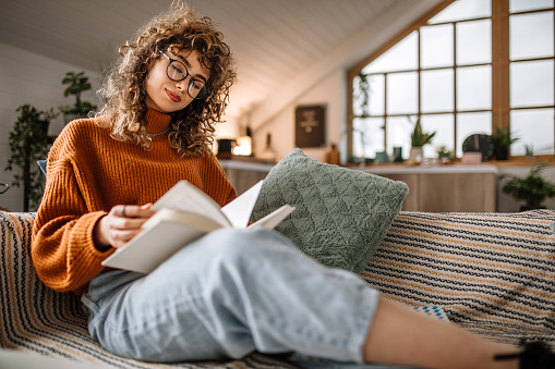 Young pretty student woman studying at coffee table, sitting on sofa in living room, reading book, writing notes