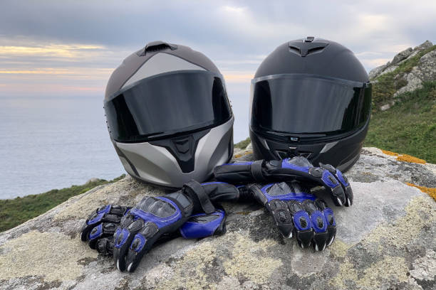 два мотоциклетных шлема с тонированными очками и кожаными мотоциклетными перчатками на скале на фоне красивого морского пейзажа - helmet helmet visor protection black стоковые фото и изображения
