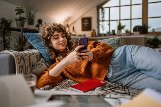 Woman using smart phone for social media laying in her couch Smiling pretty young woman, relaxes on the sofa in her living room while using her mobile smart phone for social media and surfing the internet sofa stock pictures, royalty-free photos & images