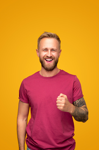 Cheerful handsome bearded young guy with tattooed arm wearing casual red t shirt looking at camera and laughing while standing against yellow background