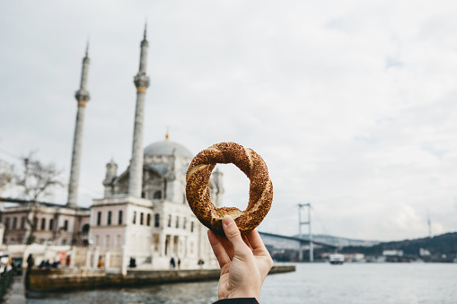 A man holds in his hand a traditional Turkish bagel called Simit against the background of the Ortakey Mosque in Istanbul in Turkey.
