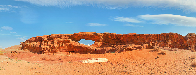Little arc or small rock window formation in Wadi Rum desert, bright sun shines on red dust and rocks, blue sky above, high resolution wide panorama