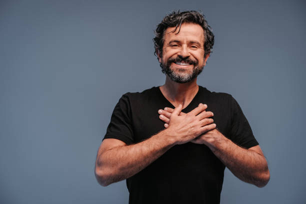 studio shot of a handsome smiling mature man with natural black and gray hair with hands on chest on grey background - self love imagens e fotografias de stock