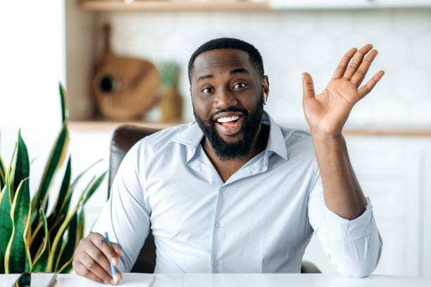istock Friendly confident successful african american young bearded man, ceo, broker or freelancer, during video conference with colleagues, taking notes, greeting with hand gesture, looks at camera, smiling 1313356846