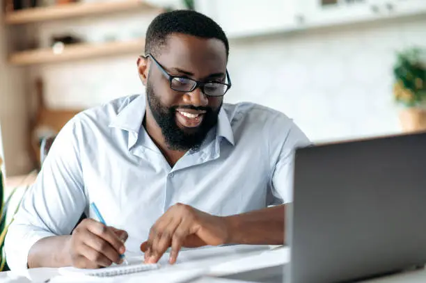 Photo of Friendly successful confident african american bearded male with glasses, broker, lawyer or manager working remotely, taking notes, talks by conference call with colleagues, distant learning, smiling
