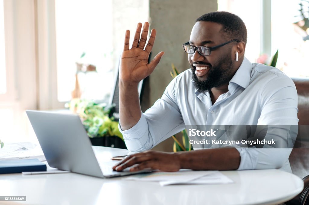 Friendly confident successful young African American businessman, manager or lawyer working at laptop, communicate with employee or customer by video conference, greeting with hand gesture, smile Working At Home Stock Photo