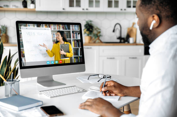 istock Online education, e-learning. African American smart confident guy sitting at home at work desk, uses computer, listening to an online lecture, taking notes, a teacher and diverse people on the screen 1313356887
