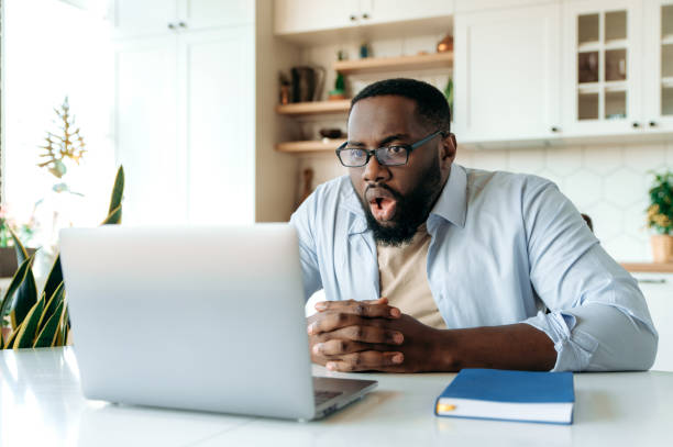 Shocked amazed African American man with glasses, freelancer, ceo or stock agent sits at his desk, looking at laptop in surprise, reading unexpected news or got big account profit Shocked amazed African American man with glasses, freelancer, ceo or stock agent sits at his desk, looking at laptop in surprise, reading unexpected news or got big account profit stock trader stock pictures, royalty-free photos & images