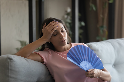 Too hot. Exhausted indian female suffer from heat in modern apartment use hand fan feel lack of air conditioning. Unhappy young mixed race female complaining on high temperature at home hard to breath