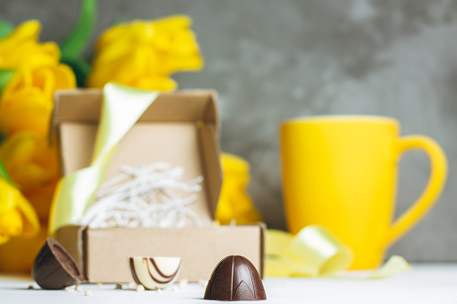 Chocolate candies in craft box, cup and bouquet of yellow tulips on white wooden surface on gray background, copy space