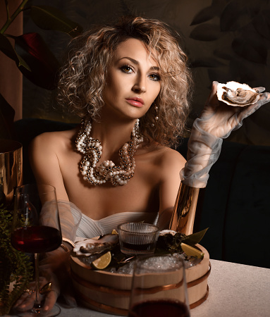 Beautiful arrogant blonde curly hair woman in seafood restaurant drinking wine and eating oysters holding one in hand going to throw over dark background