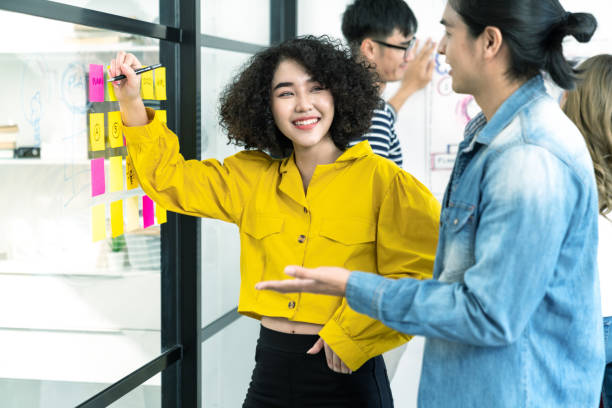 istock Asian creative woman smiling and meeting at office use post it notes to share idea and planning project. Group of employee brainstorming idea on glass wall and sticky. Brainstorming concept. 1313402166