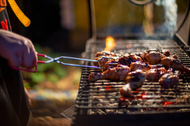 a human hand turns chicken drumsticks on a barbecue grill with grilling tongs. cooking food on an open fire in the evening. backyard party stock photo
