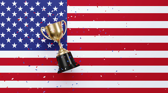 Colorful confetti falling onto a gold cup sitting over American flag background. Horizontal composition with copy space. Front view. Championship concept.