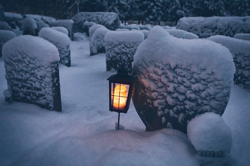 A lantern with a lit candle next to a grave at a snow covered graveyard in winter.