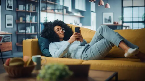 Photo of Happy Beautiful Latina Female Using Smartphone in Cozy Living Room at Home. Female Resting on Comfortable Sofa. She's Browsing the Internet and Checking Videos on Social Networks and Having Fun.
