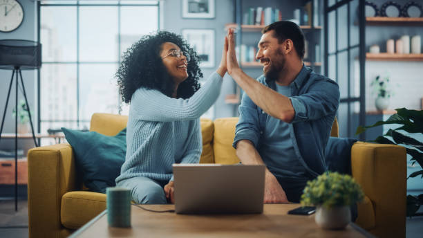 diverse multiethnische paar sitzen auf einer couch sofa in stilvollen wohnzimmer und auswahl artikel online mit laptop-computer kaufen, geben high five. freunde oder kollegen diskutieren arbeitsprojekte. - scherzare stock-fotos und bilder