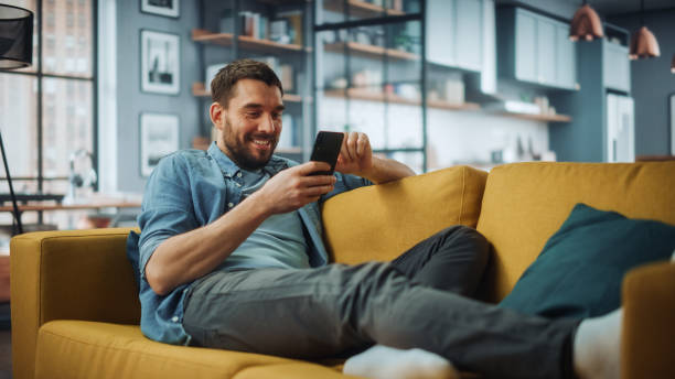 glücklich handsome kaukasischen mann mit smartphone in gemütlichen wohnzimmer zu hause. mann ruht auf bequemen sofa. er surft im internet und überprüft videos in sozialen netzwerken und hat spaß. - living room people joy happiness stock-fotos und bilder